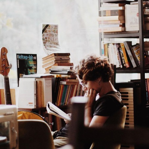 woman, surrounded by books, sits and reads in front of a picture window